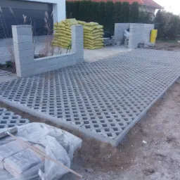 Układanie betonowej ażurowej kostki brukowej na podjeździe, widoczne worki z żółtym piaskiem, częściowo zbudowane ogrodzenie z betonowych bloczków i ubijarka wibracyjna.