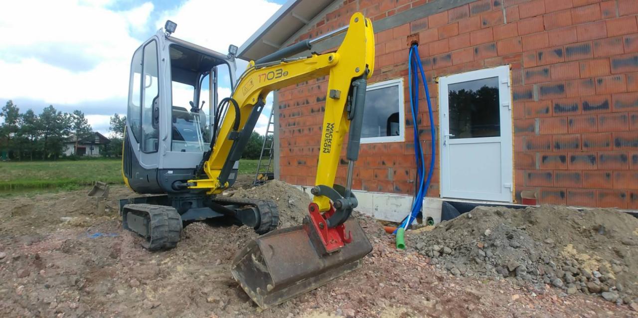 Żółto-szara minikoparka Wacker Neuson 1703e na gąsienicach, z łyżką skierowaną w stronę budynku z czerwonej cegły, w tle zielony teren z domem i drzewami, widoczne niebieskie rury wchodzące...