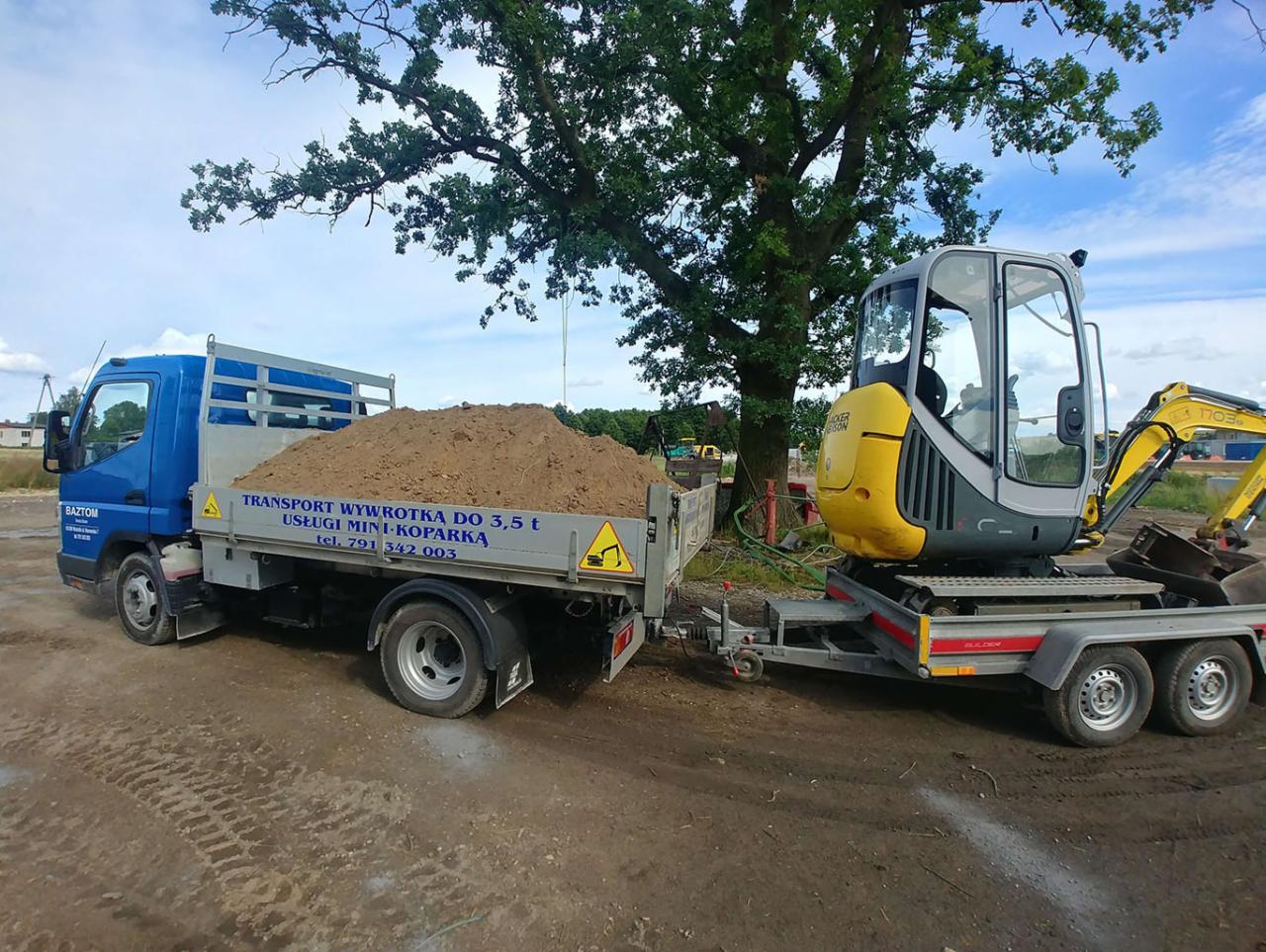 Niebieska wywrotka z ładunkiem ziemi oraz żółto-szara mini-koparka na przyczepie, zaparkowane na ziemistym terenie pod rozłożystym drzewem, w tle inne maszyny budowlane.