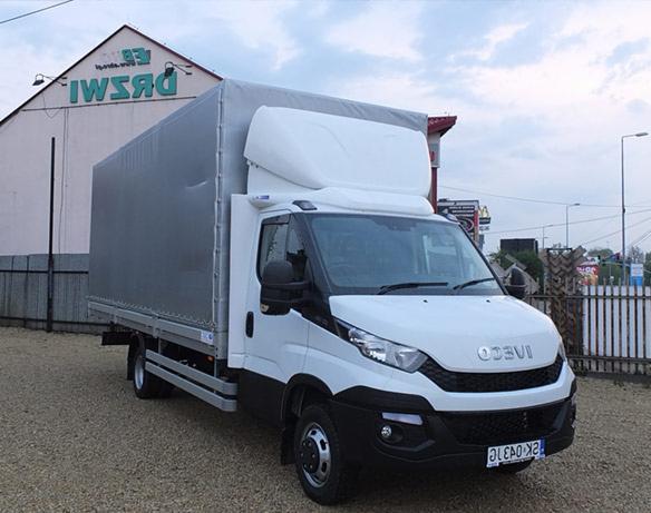 Biały samochód dostawczy Iveco z szarą plandeką na tle budynku z zielonym napisem, sfotografowany z lekkiego dołu.