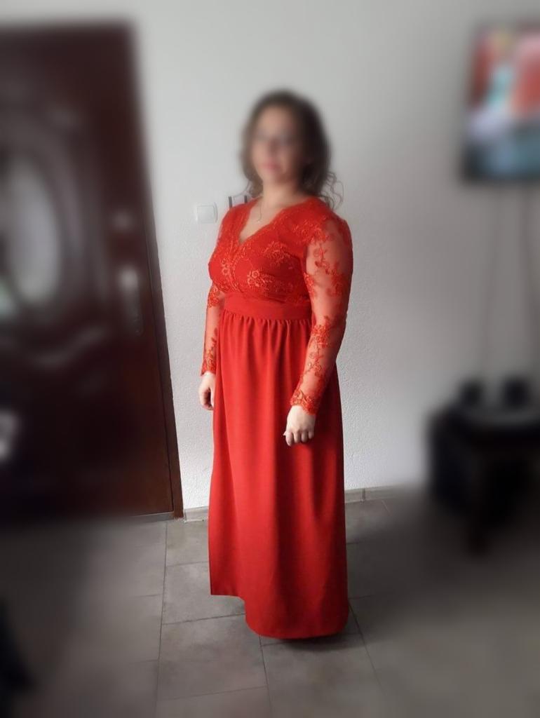 Kobieta pozuje w długiej, czerwonej sukni z koronkowymi rękawami i dekoltem w kształcie litery V, stojąc na tle białej ściany i brązowych drzwi.