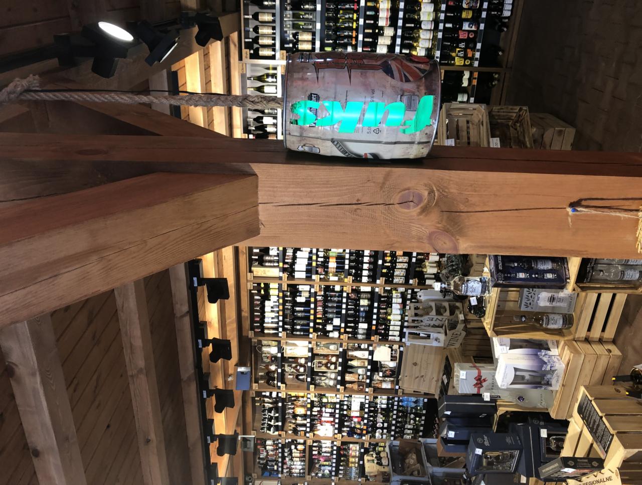 Wnętrze sklepu z alkoholem w Bytomiu, widok na drewnianą konstrukcję sufitu i regały wypełnione butelkami wina i innych trunków, oświetlone punktowymi lampami.