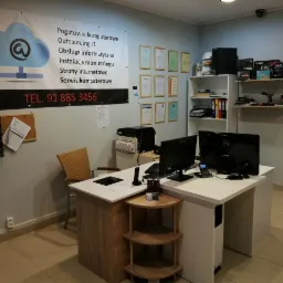 Wnętrze biura firmy IT z biurkiem, dwoma monitorami, bannerem reklamowym z usługami, półką z komponentami komputerowymi i certyfikatami na ścianie.