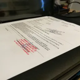 Dokument z pieczątką firmy AdeNET Wsparcie IT, serwis komputerowy, leżący na czarnym segregatorze, w tle monitor komputera.