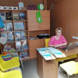 Kobieta w różowej bluzce siedzi przy biurku z laptopem, na którym leżą ulotki z motywem plaży; w tle regał z broszurami, książkami i balonem reklamowym.