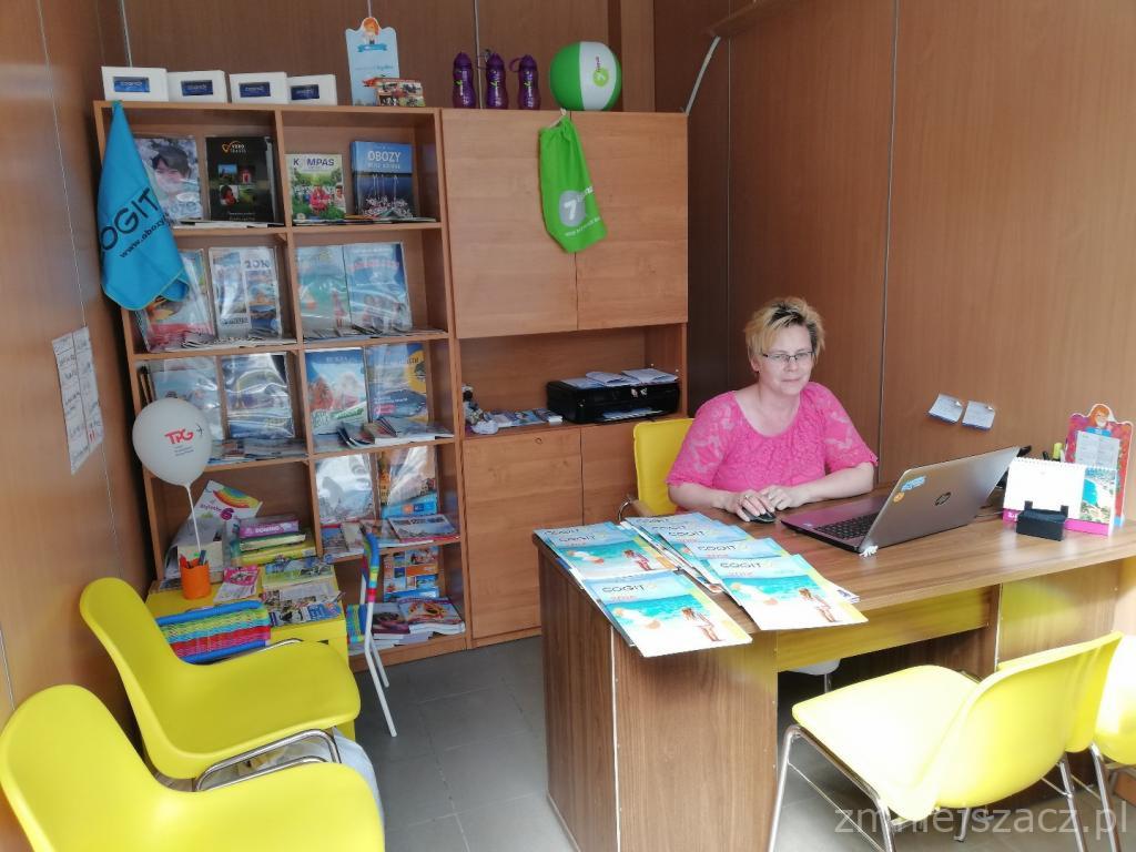 Kobieta w różowej bluzce siedzi przy biurku z laptopem, na którym leżą ulotki z motywem plaży; w tle regał z broszurami, książkami i balonem reklamowym.