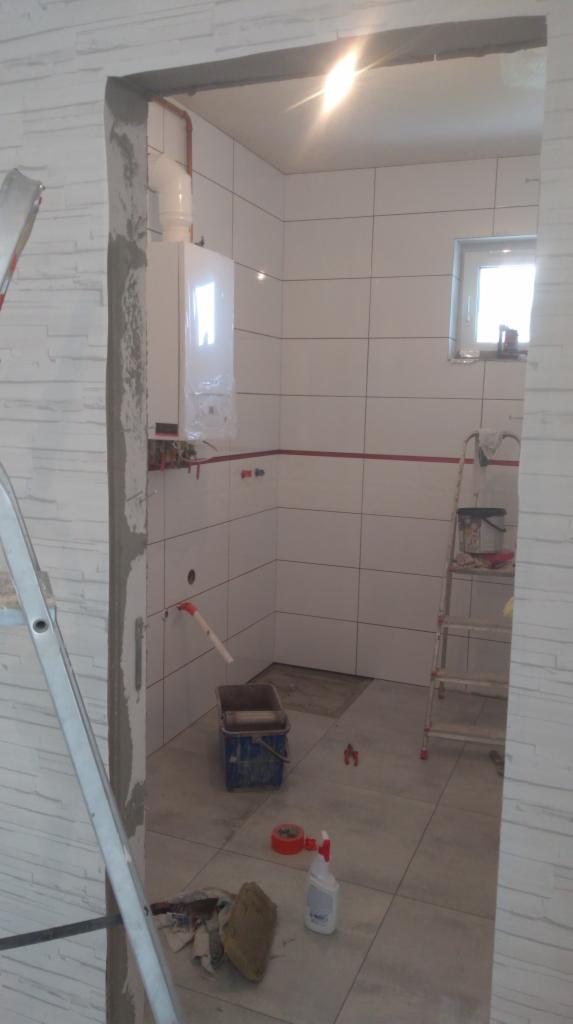 Wnętrze łazienki w trakcie prac wykończeniowych: białe płytki ścienne z czerwoną fugą, drabina z narzędziami, bojler, fragment ściany z dekoracyjną fakturą, niebieskie wiadro i akcesoria na podłodze.