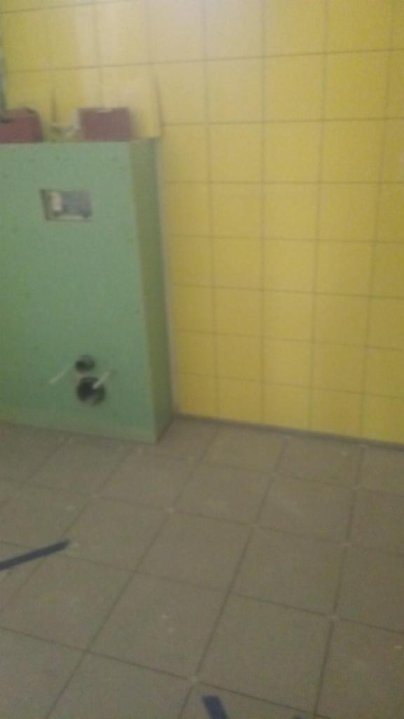 Żółte płytki ścienne i szare płytki podłogowe w łazience, z zieloną obudową instalacji podtynkowej. Widoczne rury wystające ze ściany.