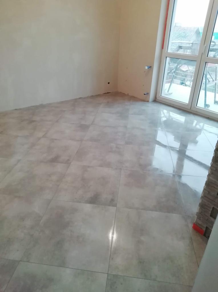 Jasne, kwadratowe płytki podłogowe w pustym pokoju z dużym oknem balkonowym i przygotowanymi instalacjami na ścianie.