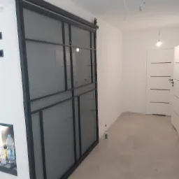 Szklane drzwi przesuwne w czarnej metalowej ramie zamontowane na szynie w jasnym wnętrzu, widoczne elementy mocujące.