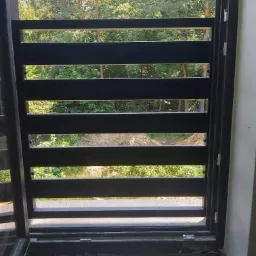 Czarna, pozioma balustrada balkonowa z widokiem na zieleń drzew w tle, zamontowana na mokrej podłodze balkonu.