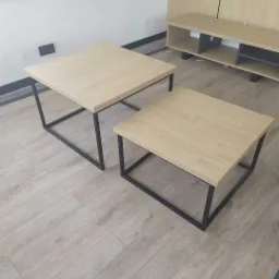 Dwa stoliki kawowe o kwadratowych blatach z jasnego drewna i czarnych, metalowych nogach o prostej konstrukcji, stojące na podłodze z paneli w odcieniach szarości, w tle widoczna szafka RTV...