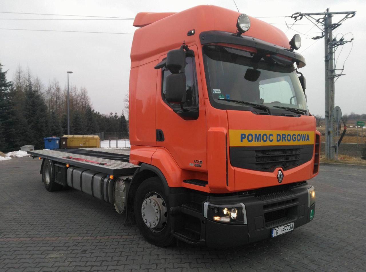 Pomarańczowa laweta Renault z napisem 'POMOC DROGOWA' na tle szarej kostki brukowej i drzew bez liści