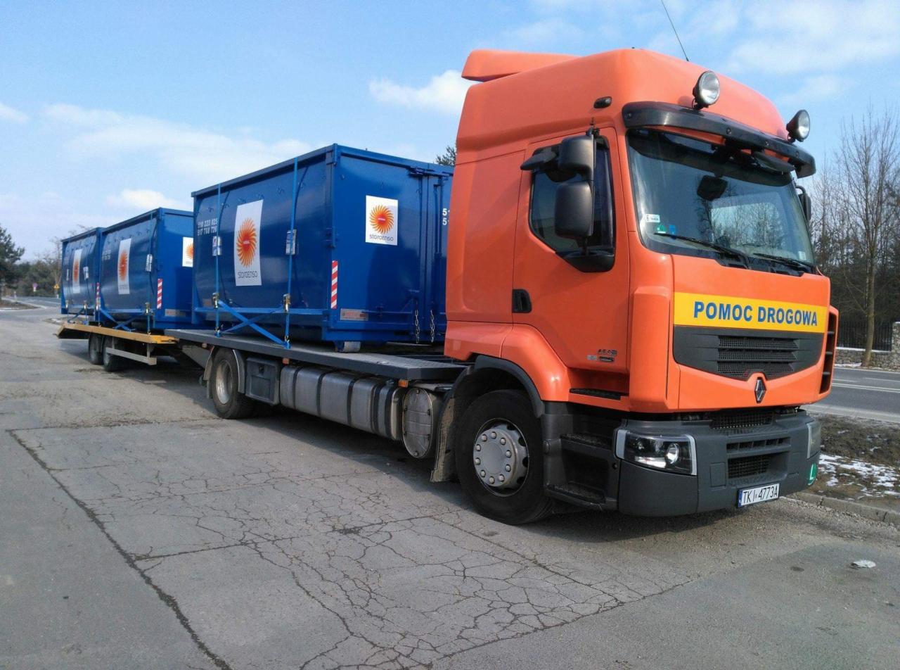 Pomarańczowa ciężarówka z napisem 'POMOC DROGOWA' przewozi na platformie dwa niebieskie kontenery z logo firmy.