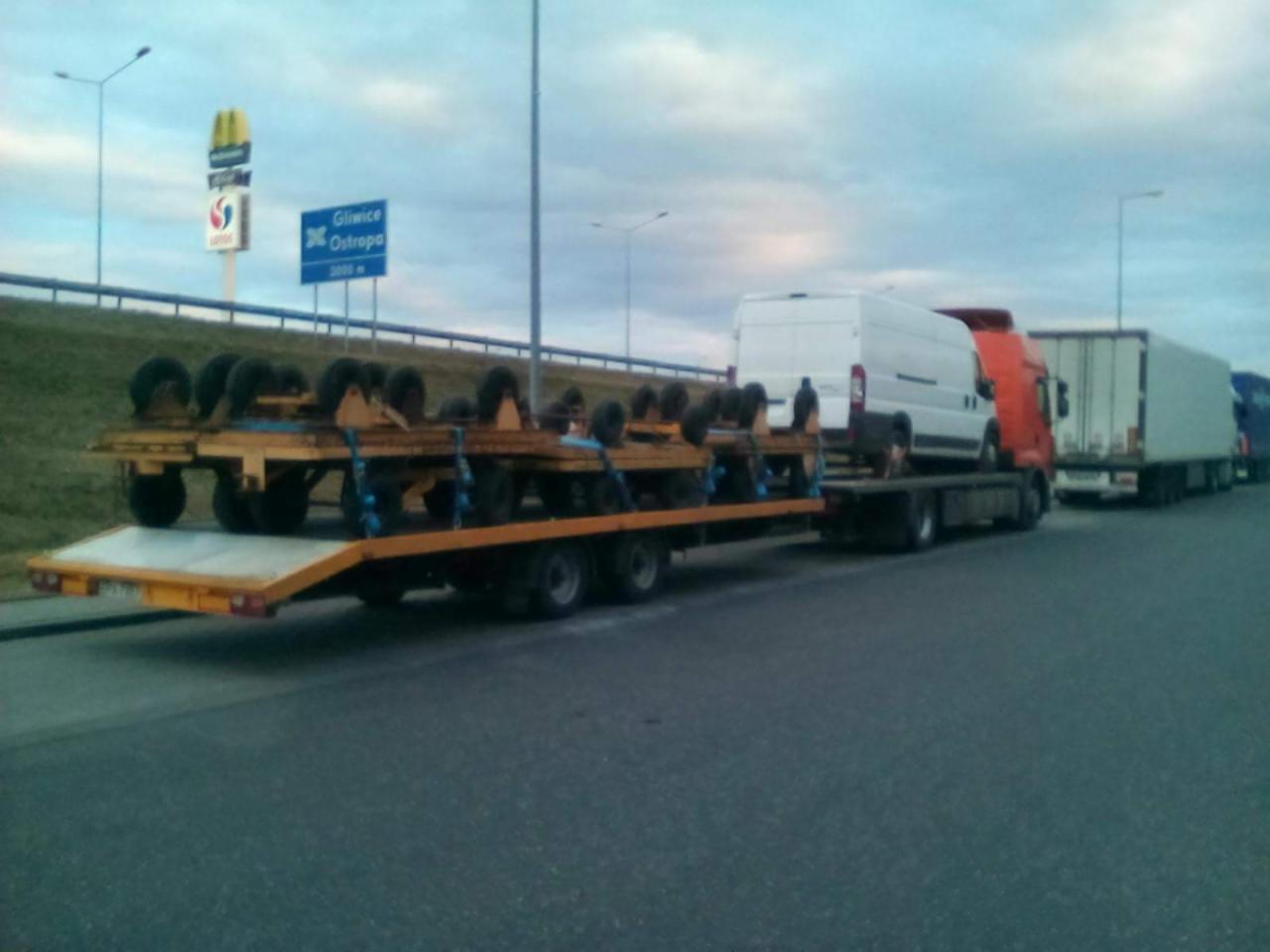 Żółta platforma z kołami, biały bus na czerwonej ciężarówce i biała naczepa na autostradzie z drogowskazem Gliwice-Ostroppa w tle.