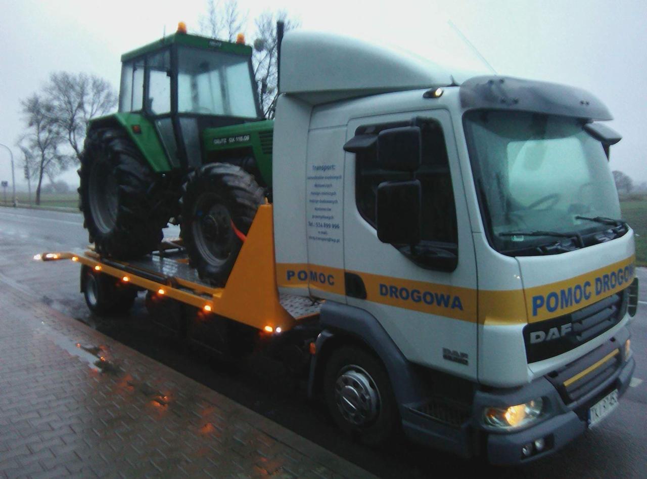 Laweta transportuje zielony ciągnik Deutz-Fahr na mokrej, asfaltowej drodze w pochmurny dzień.