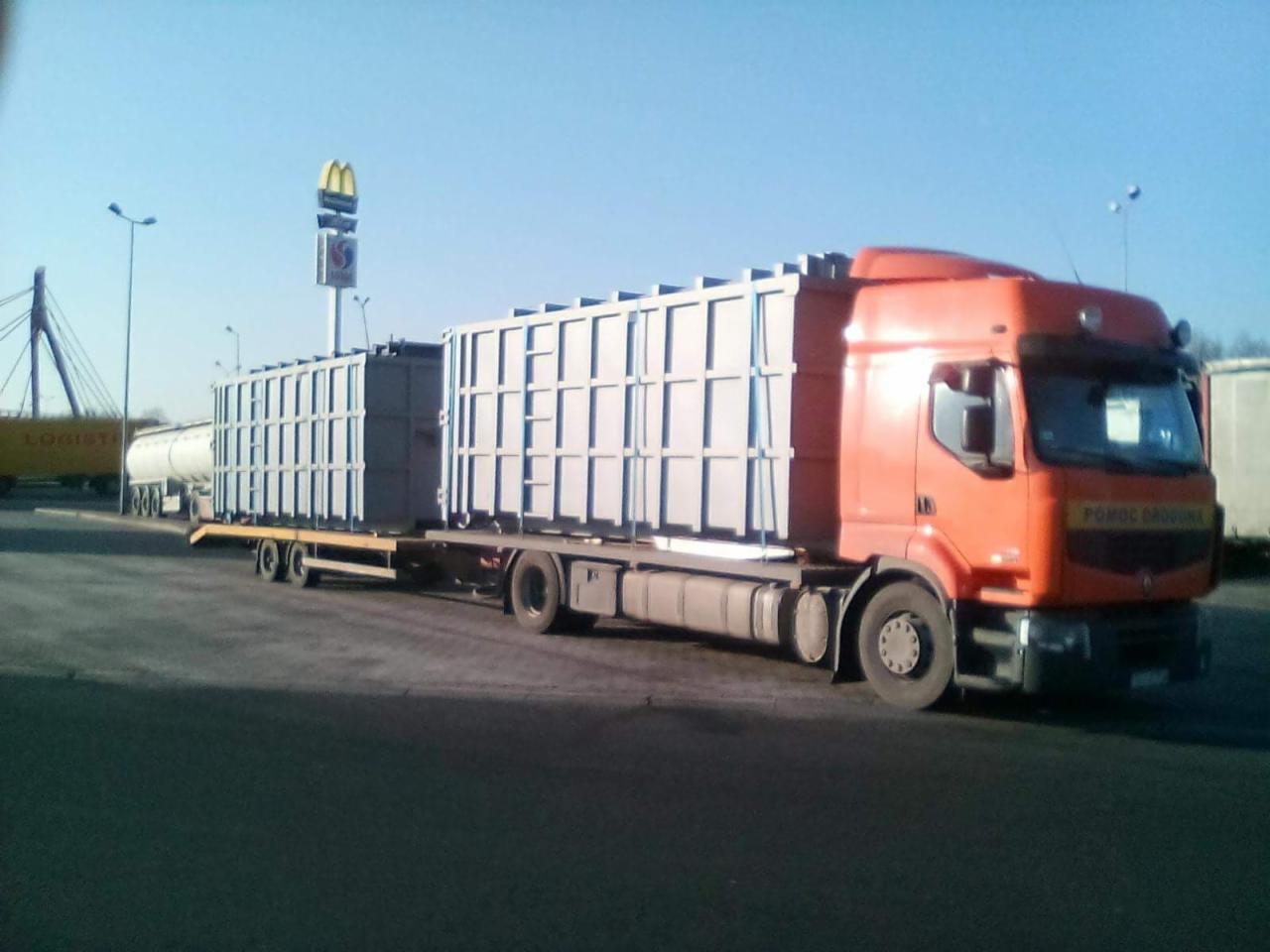 Pomarańczowa ciężarówka z naczepą przewożąca dwa duże, szare kontenery z kratownicową konstrukcją, zaparkowana na parkingu z widocznym w tle znakiem McDonald's i wiaduktem.