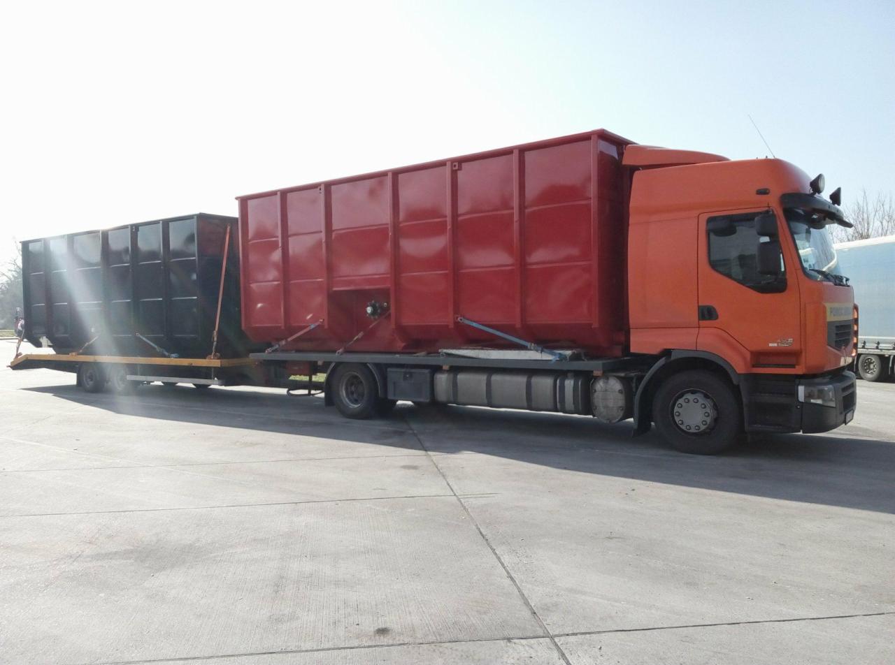 Pomarańczowa ciężarówka Renault z czerwoną zabudową kontenerową oraz czarnym kontenerem na przyczepie, zabezpieczone pasami transportowymi, na betonowym placu w słoneczny dzień.