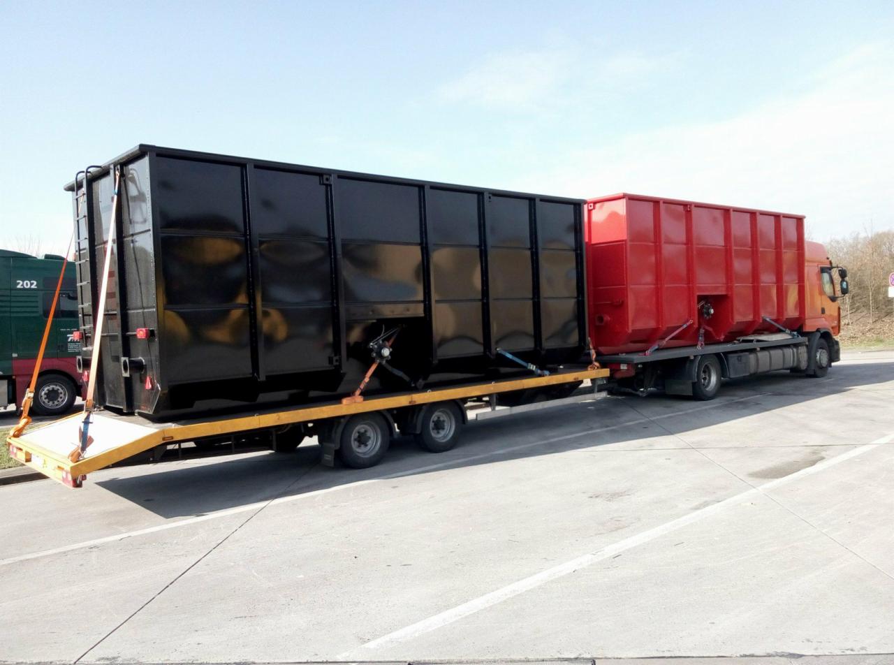 Ciężarówka z pomarańczową kabiną przewozi na niskopodwoziowej przyczepie dwa duże kontenery: czarny i czerwony zabezpieczone pasami transportowymi.