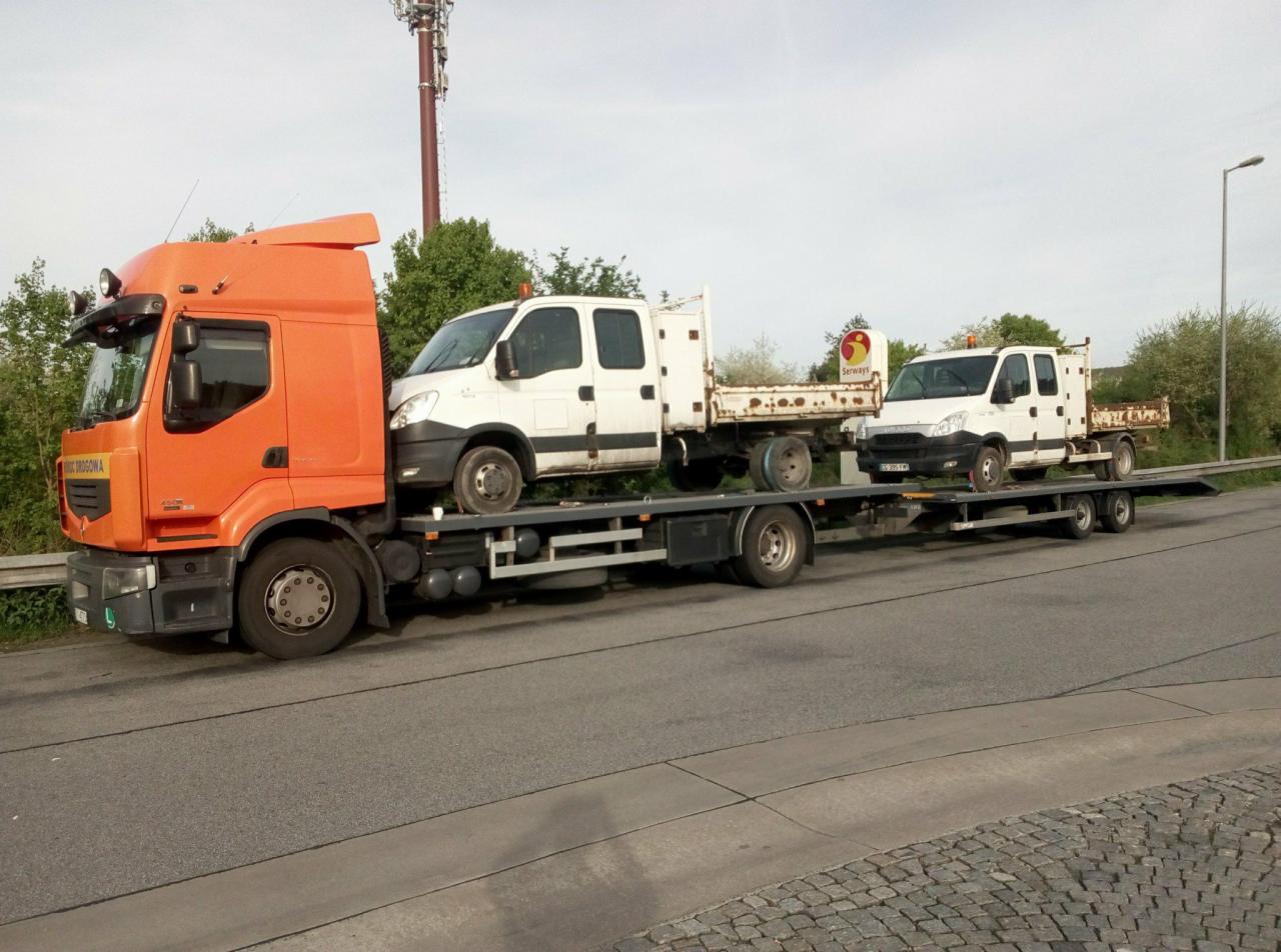 Pomarańczowa ciężarówka z lawetą przewożąca dwa białe samochody dostawcze na tle zieleni i słupa telekomunikacyjnego.