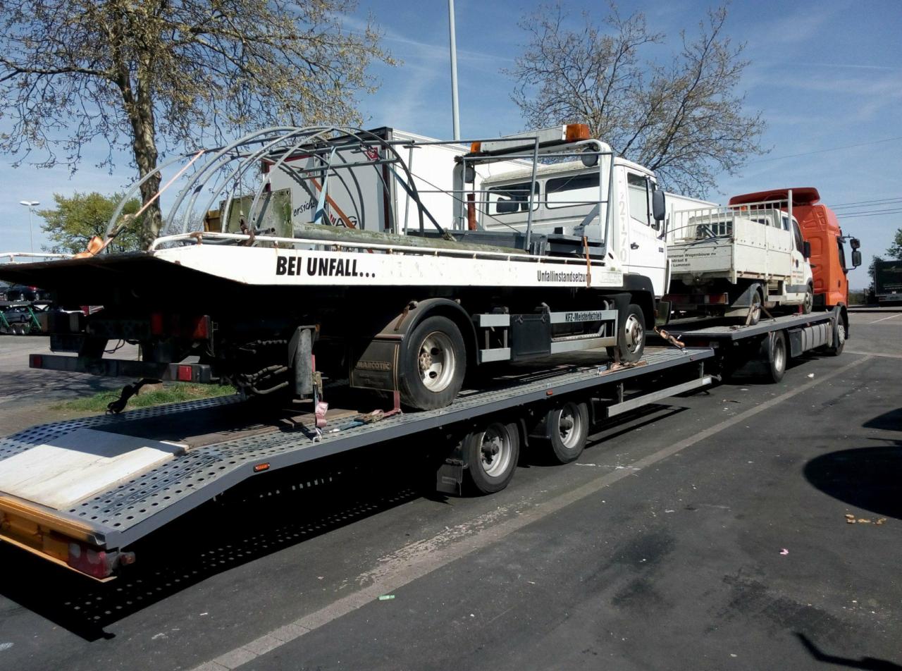 Laweta przewozi białą ciężarówkę z napisem 'BEI UNFALL...' oraz małą białą ciężarówkę na platformie, widoczne drzewa w tle.