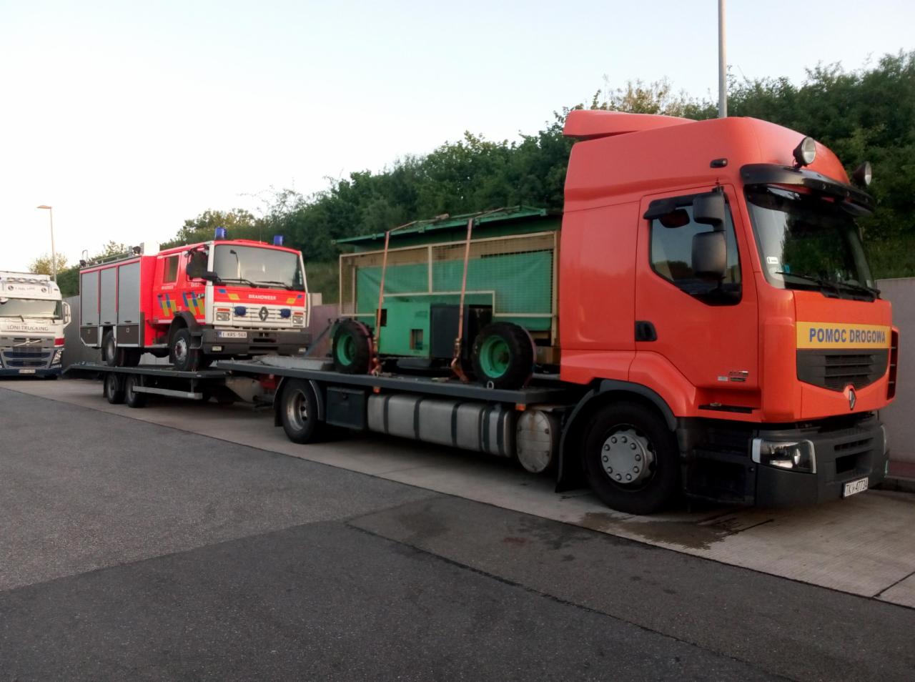 Pomarańczowa ciężarówka z napisem 'Pomoc Drogowa' przewozi na platformie zielony sprzęt rolniczy i czerwony wóz strażacki z niebieskimi światłami.