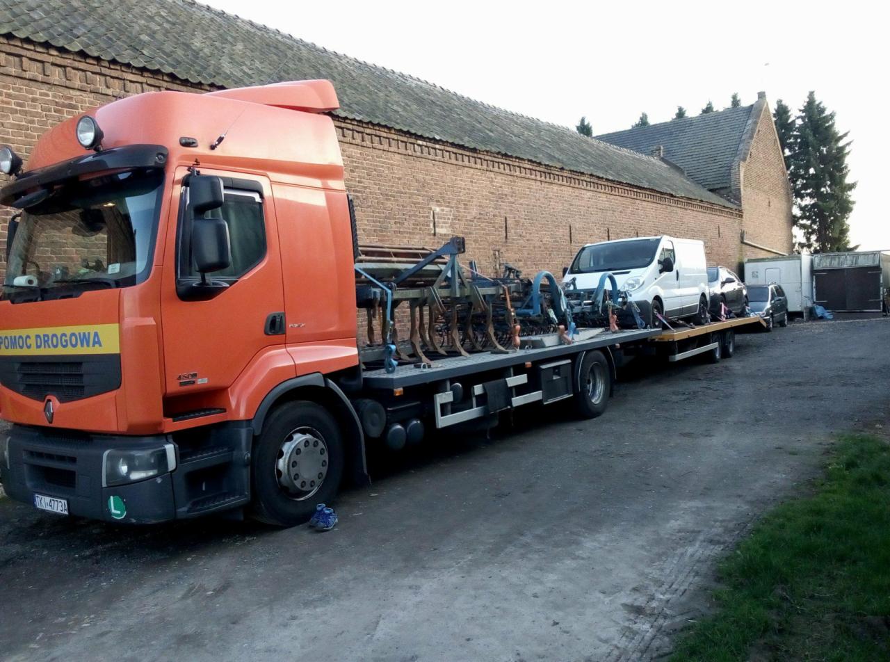 Pomarańczowa laweta z napisem 'Pomoc Drogowa' przewozi biały bus i dwa samochody osobowe na tle ceglanego budynku gospodarczego.