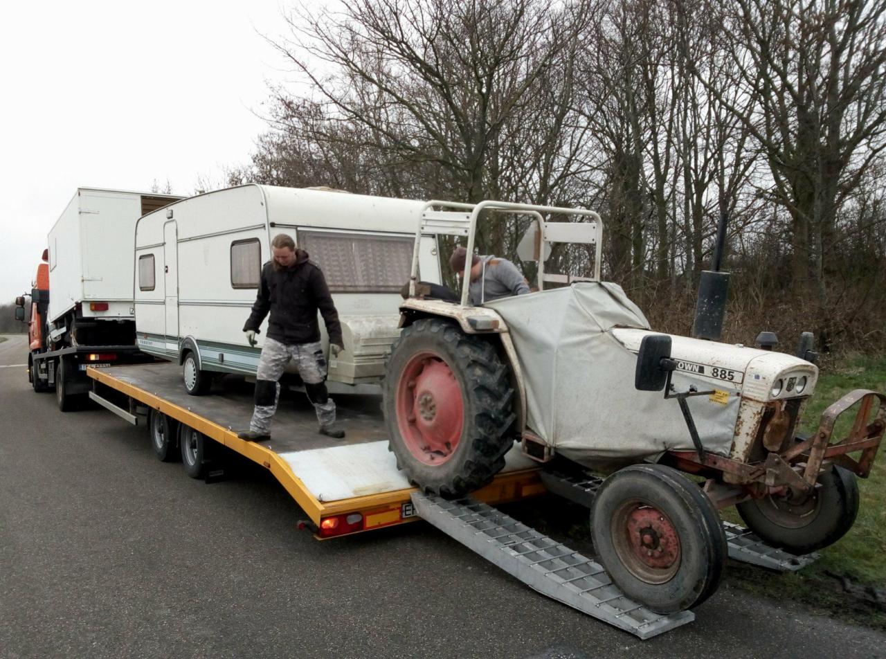 Tractor and caravan being loaded onto a yellow flatbed truck using ramps, with two men assisting; trees in the background.