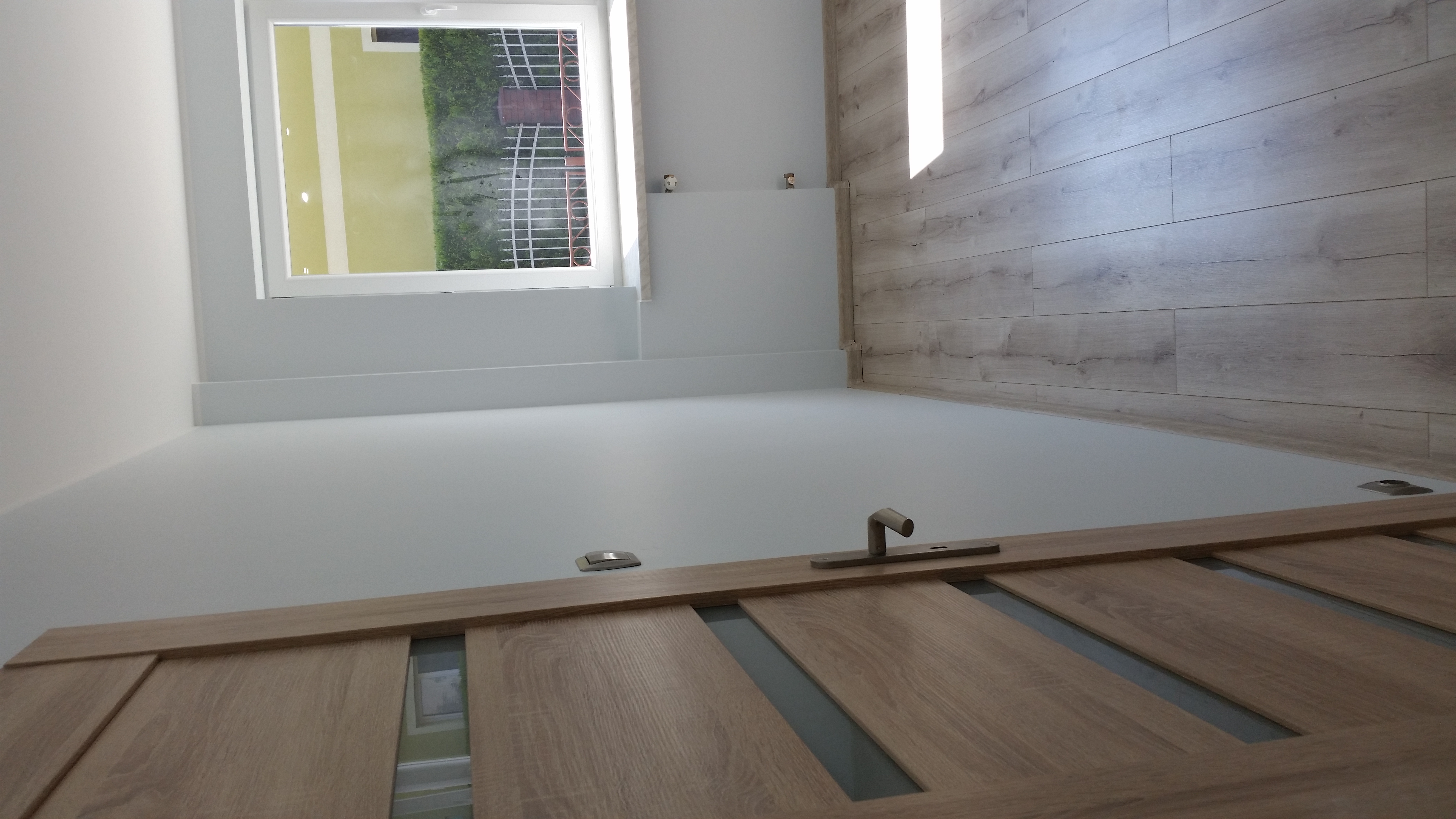 Wnętrze po remoncie: gładka, biała ściana, jasne panele podłogowe o strukturze drewna i fragment drewnianych drzwi z przeszkleniami, widok z dołu.