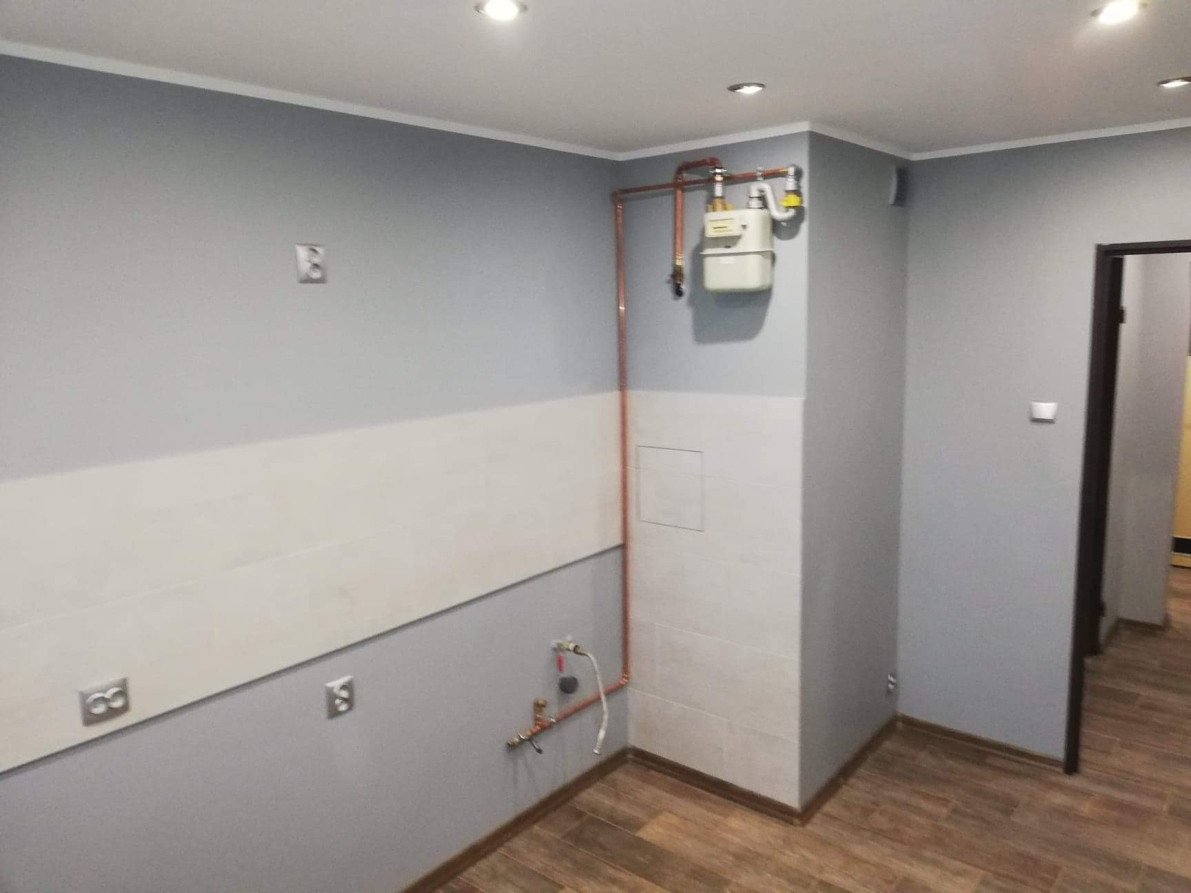 Świeżo pomalowane na szaro pomieszczenie z częściowo wykończoną białą lamperią, brązowymi panelami podłogowymi i widoczną instalacją gazową z licznikiem.