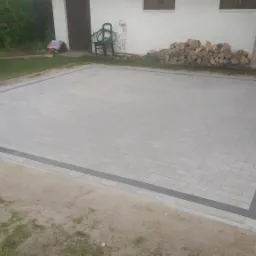 Nowo ułożona, prostokątna powierzchnia z szarej kostki brukowej z obramowaniem z ciemniejszych kostek, widok z góry, w tle budynek gospodarczy, ułożone drewno i zieleń.