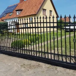 Czarna, metalowa brama przesuwna z dekoracyjnymi elementami w kształcie koszyczków, zamontowana przed domem z panelami słonecznymi na dachu.