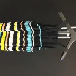 Plisowana sukienka w czarno-pastelowe paski, wisząca na wieszaku na tle ciemnej ściany, widoczny fragment metalowego stojaka na ubrania.