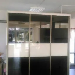 Trzydrzwiowa szafa przesuwna z czarno-białymi panelami lakierowanymi, widoczna w jasnym wnętrzu biurowym z oświetleniem LED na suficie.