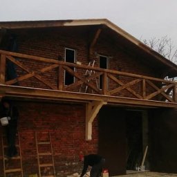 Felc - Budynek z cegły z drewnianym balkonem w trakcie budowy. Widoczne rusztowania i osoby pracujące przy wykończeniu. Elementy drewniane w surowym stanie.