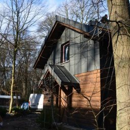 Felc - Dom z ciemną, metalową elewacją i dachem, otoczony drzewami. Widoczny fragment rusztowania i pracownik na dachu. Nietypowe połączenie materiałów elewacyjnych.