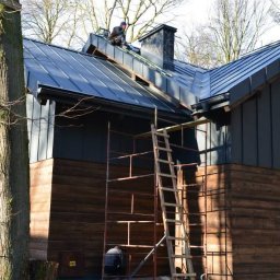 Felc - Dom z ciemną, metalową blachą na dachu i drewnianą elewacją. Na dachu pracuje dekarz, widoczne rusztowania i komin. Ujęcie z dołu, drzewo na pierwszym planie.