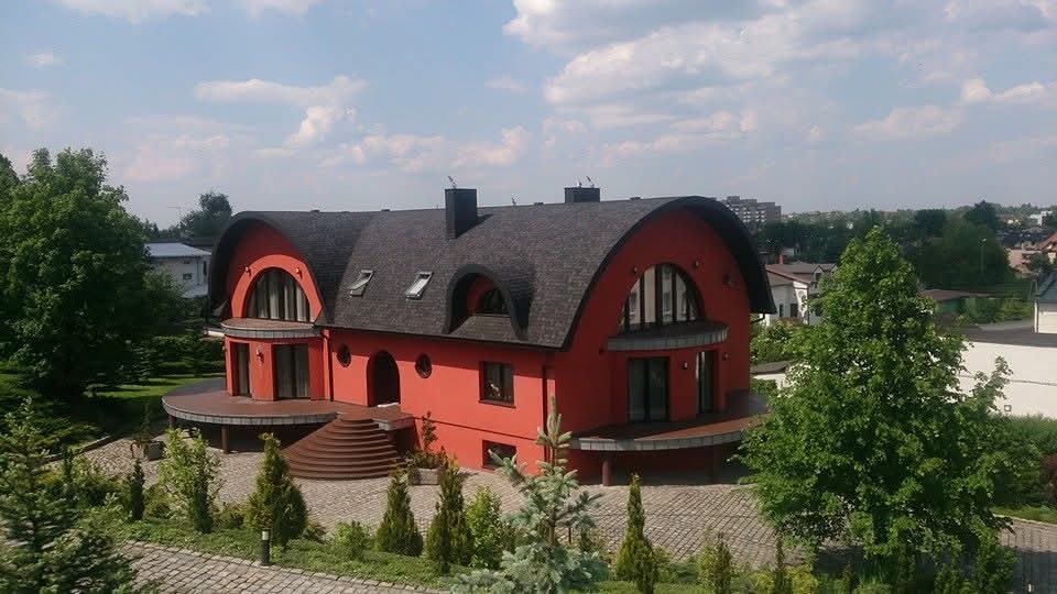 Czerwony dom z ciemnoszarym dachem o nietypowym kształcie, widok z góry, otoczony zielenią i brukowanym podjazdem, w tle zabudowa miejska.