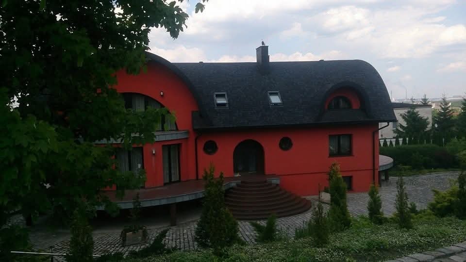 Czerwony dom z ciemnym, asymetrycznym dachem krytym gontem bitumicznym, widoczne okna dachowe i komin. Ujęcie z poziomu ogrodu, częściowo zasłonięte drzewem.