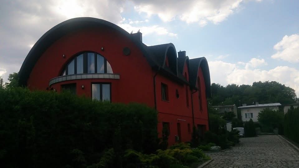 Czerwony dom z nietypowym, łukowym dachem krytym czarną dachówką, widoczny zza zieleni. Na dachu siedzą dwa ptaki. Niebo częściowo zachmurzone.