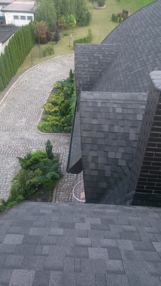 Szary dach z gontów bitumicznych, widok z góry na fragment dachu z kominem i rynną, na tle brukowanej ścieżki i zieleni.