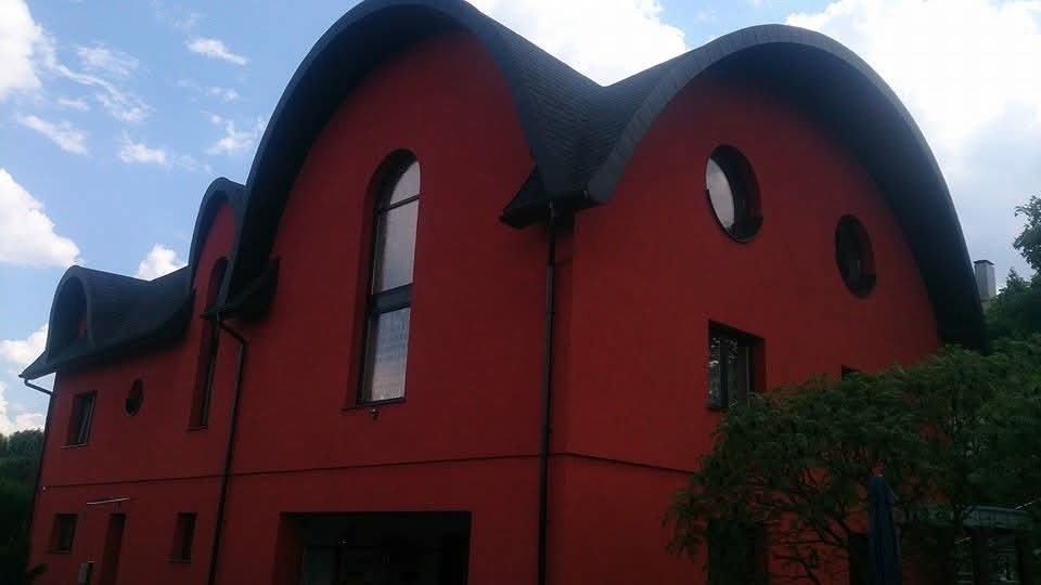 Czerwony budynek z nietypowym, czarnym dachem o zaokrąglonych kształtach i owalnymi oknami, widziany z dołu pod kątem. Widoczne rynny i fragment zieleni.
