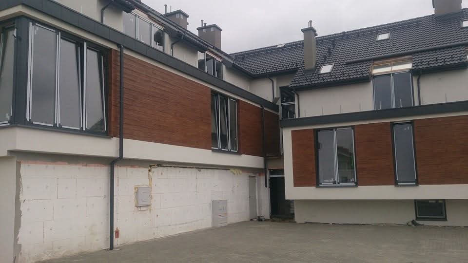 Dwupiętrowy budynek z ciemną dachówką i drewnianą elewacją. Widoczne okna z szarymi ramami i fragment dachu z kominami. Część budynku w trakcie wykończenia.