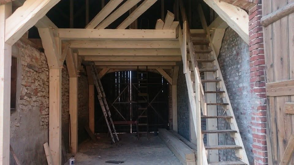Wnętrze stodoły z nową, drewnianą konstrukcją więźby dachowej i schodami, surowe ściany z kamienia i cegły, widoczne drabiny i materiały budowlane.