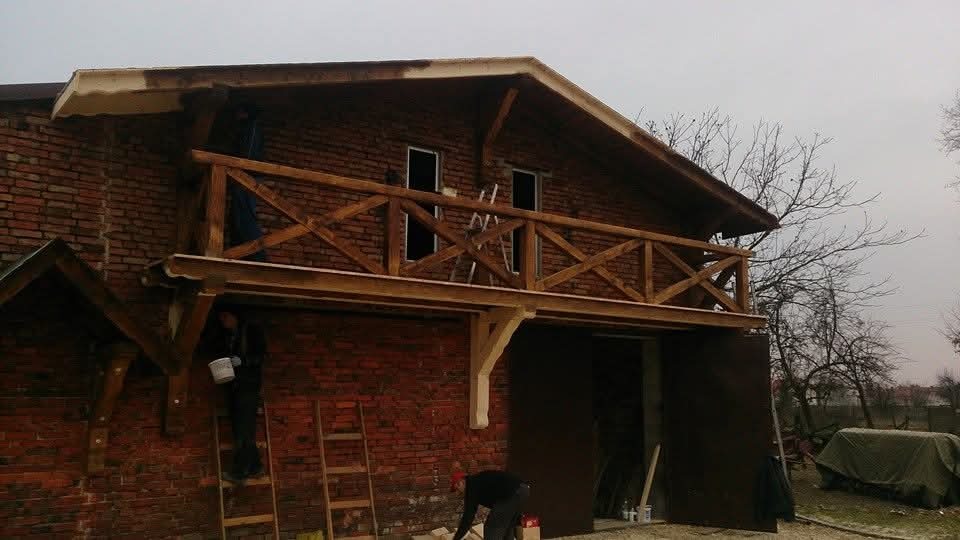 Budynek z cegły z drewnianym balkonem w trakcie budowy. Widoczne rusztowania i osoby pracujące przy wykończeniu. Elementy drewniane w surowym stanie.