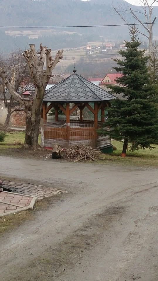 Drewniana altana z szarym dachem krytym dachówką bitumiczną na tle górskiego krajobrazu. Widoczne drzewa i ścieżka.