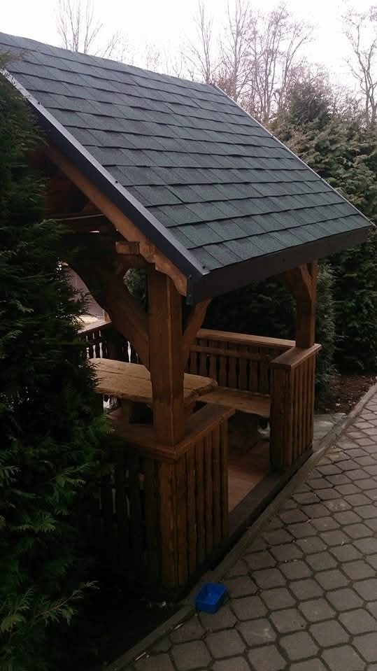 Drewniana altana z dachem pokrytym gontem bitumicznym, otoczona zielenią. Widoczny stół i ławki wewnątrz konstrukcji. Na ziemi leży niebieski pojemnik.