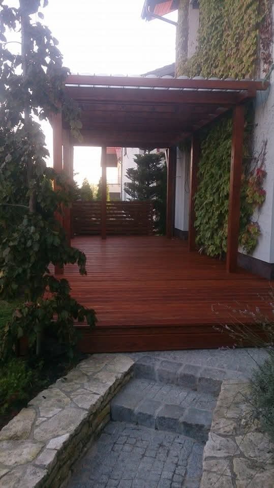 Drewniana pergola z tarasem, zadaszona konstrukcja z widocznymi deskami podłogowymi i elementami roślinnymi na ścianie budynku. Kamienne schody prowadzą na taras.