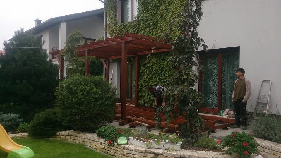 Montaż drewnianej pergoli przy domu z elewacją porośniętą bluszczem. Widoczni dwaj mężczyźni podczas pracy, narzędzia i elementy konstrukcyjne.