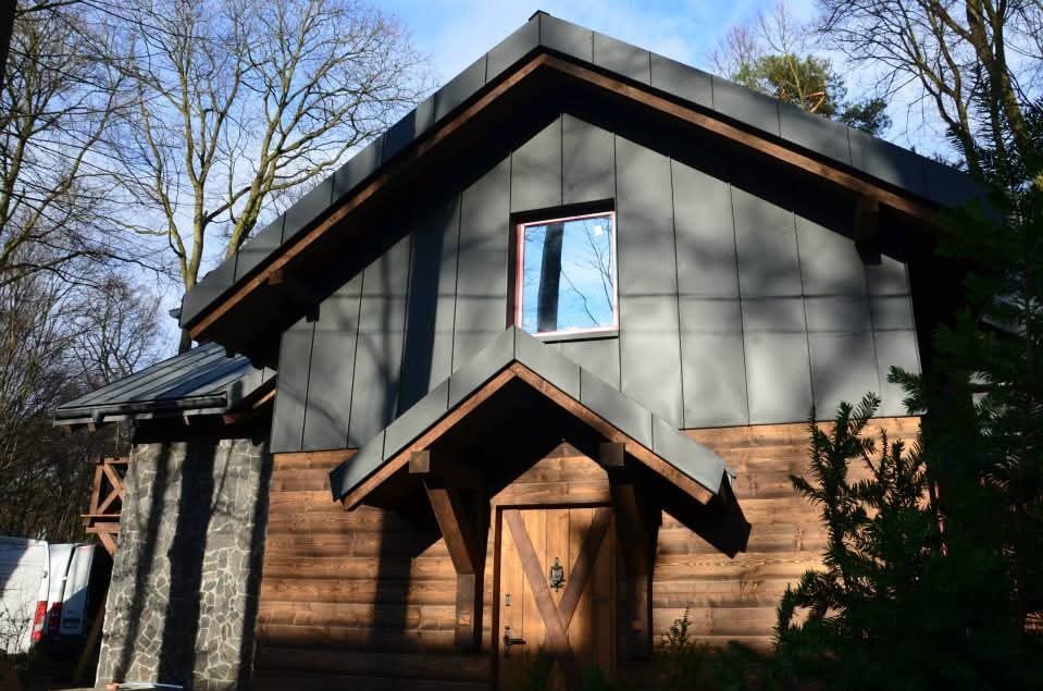 Dom z szarą blachą na dachu i drewnianą elewacją, widok z dołu, okno z odbiciem nieba, drzewa w tle, kamienna kolumna po lewej stronie.